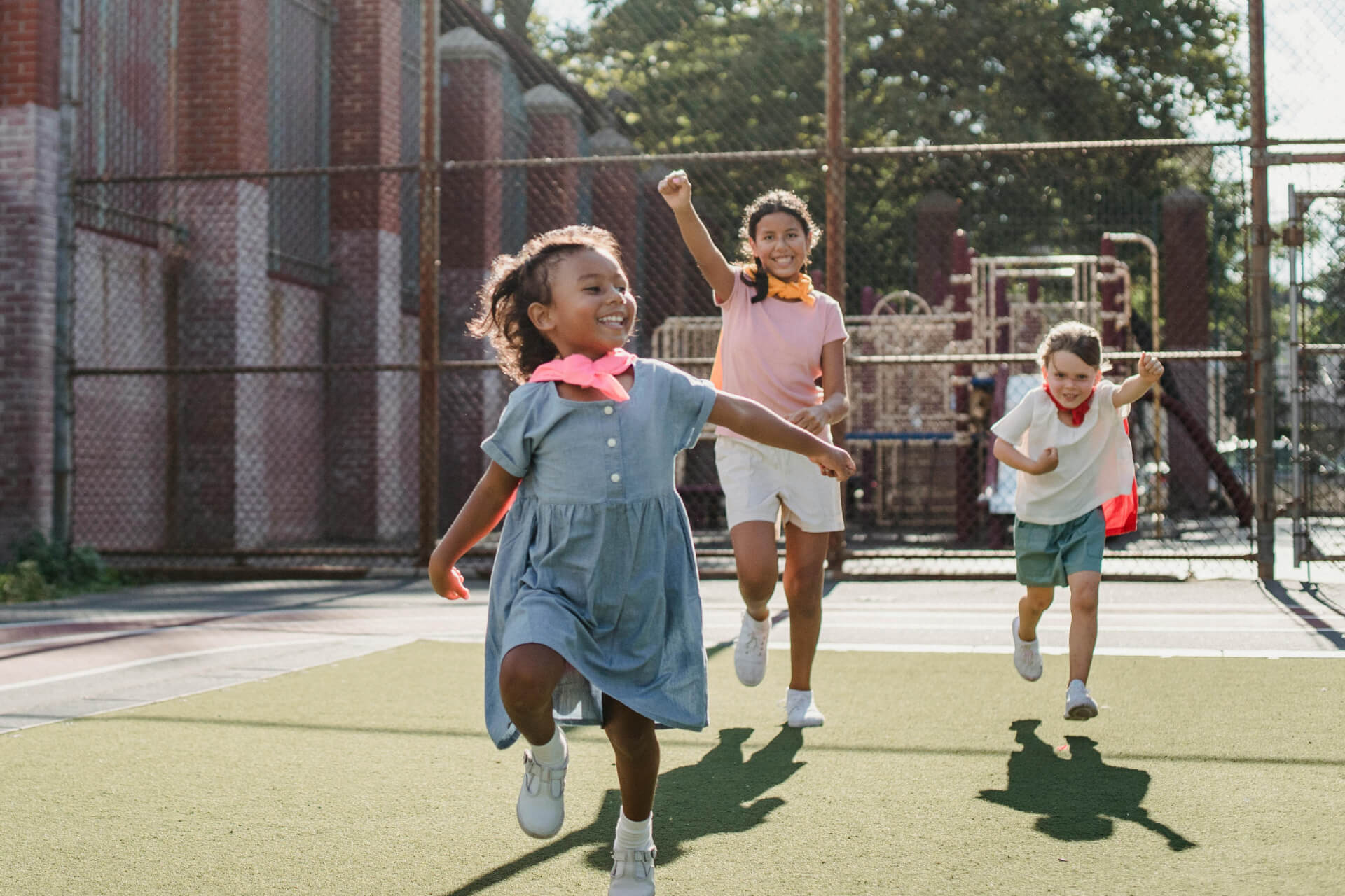 Three children wearing casual clothes run and play on an outdoor 
            sports court with a playground and fence in the background.