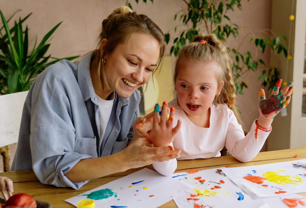 A woman and a young girl with Down syndrome smile while painting with their hands at a table, surrounded by colorful paint and paper.