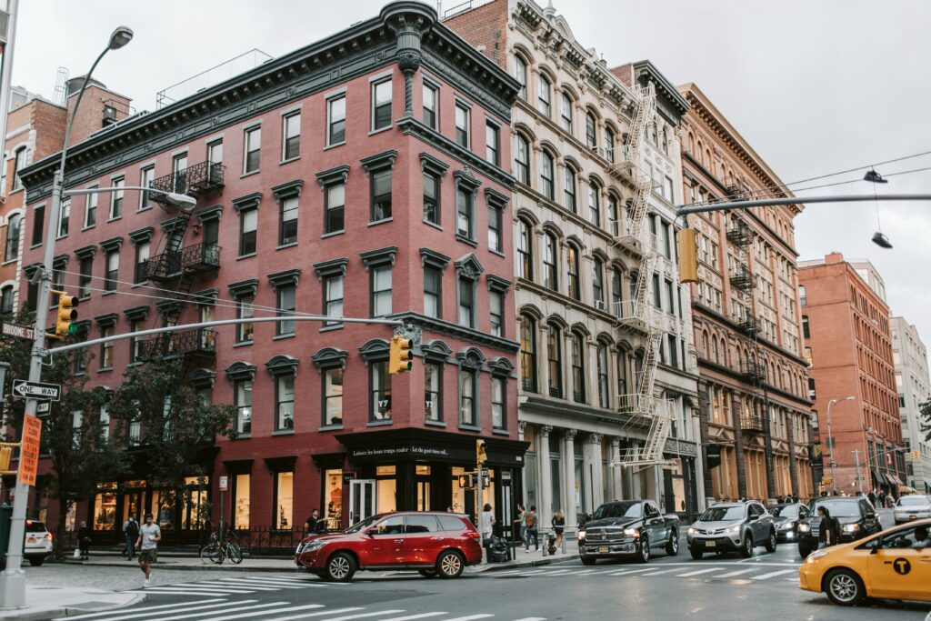 A busy city intersection with red and beige historic buildings, cars and a yellow taxi waiting at the traffic light, pedestrians on the sidewalks, and trees lining the street on a cloudy day.