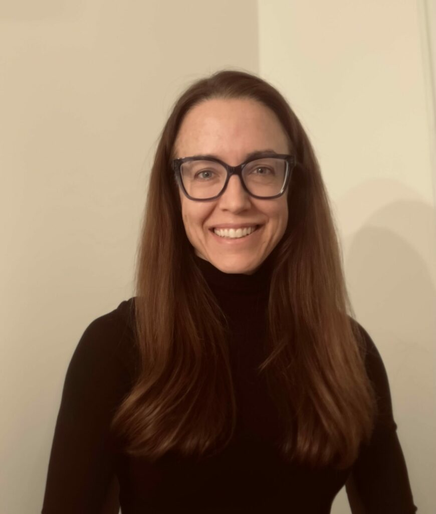 A woman with long brown hair, wearing glasses and a black turtleneck, smiles at the camera against a plain, light-colored background.